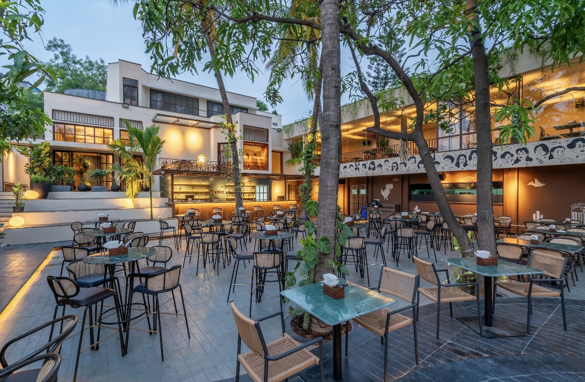 Empty tables and chairs across the open yard in daylight, trees overhead.