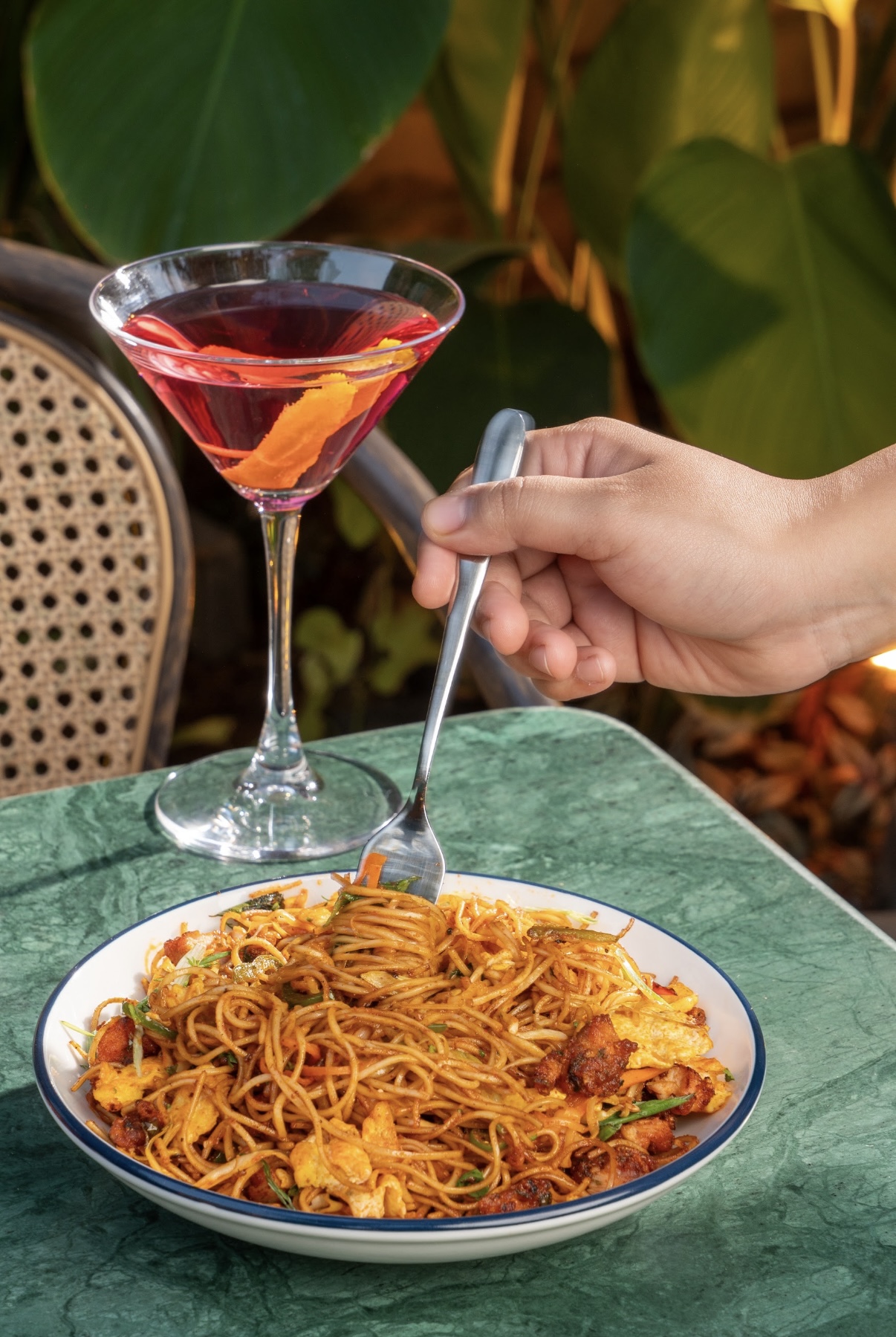 A hand twirling pasta beside a rose-coloured cocktail on a green marble table.