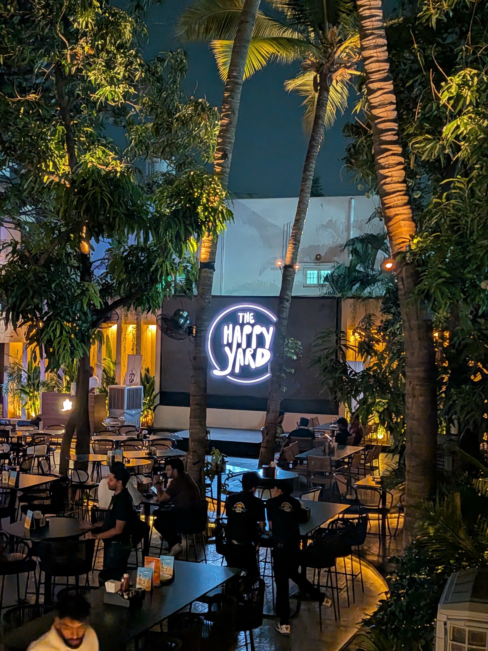 The yard after dark, palms lit from below, the logo glowing on the wall.