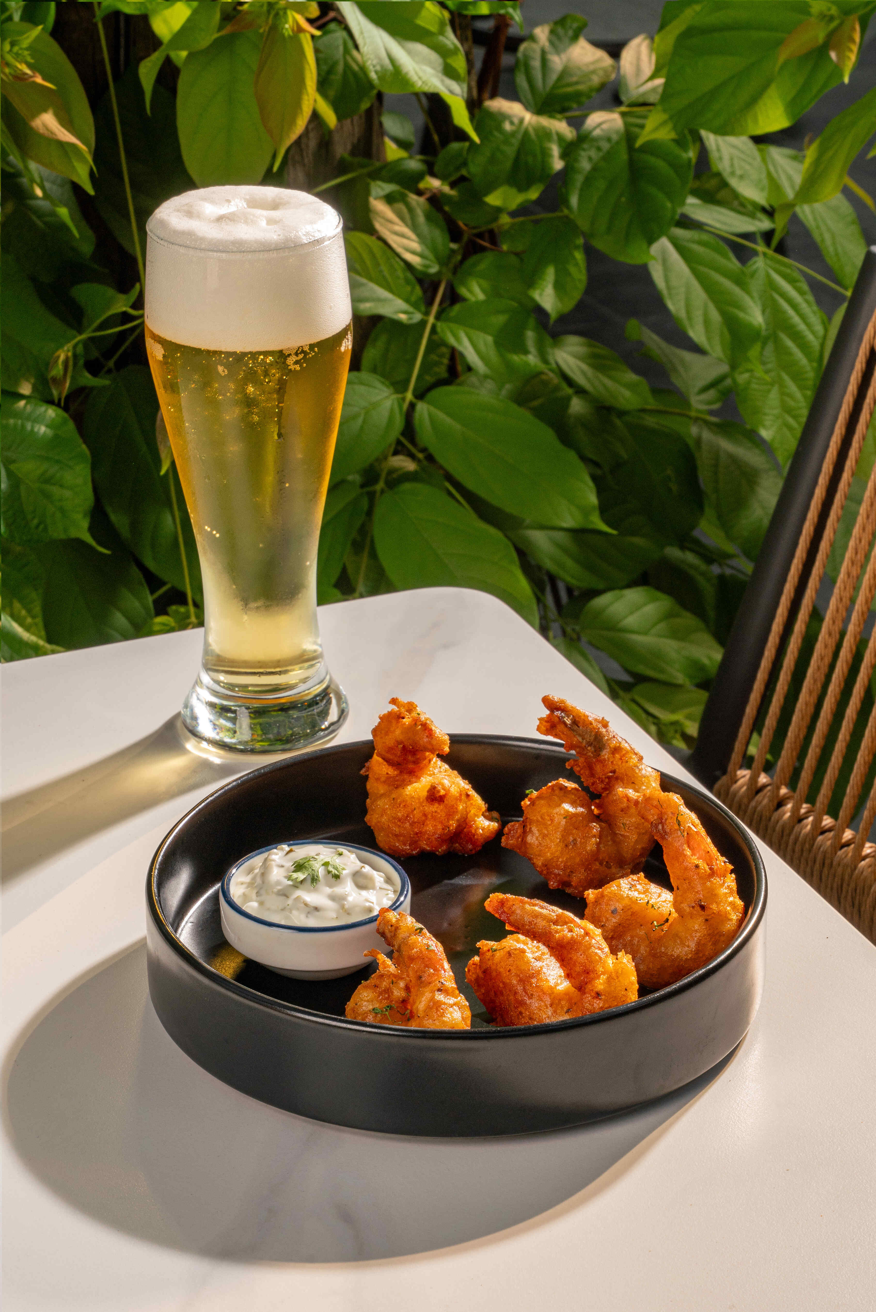 A tall beer beside battered prawns and a dip, greenery behind.